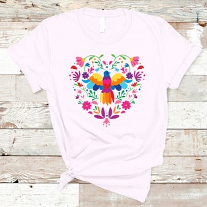 May include: A white t-shirt with a colorful heart-shaped design featuring a bird and flowers. The design is in a traditional Mexican folk art style.