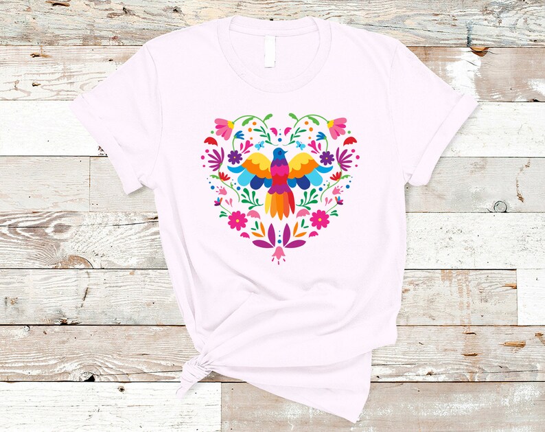 May include: A white t-shirt with a colorful heart-shaped design featuring a bird and flowers. The design is in a traditional Mexican folk art style.