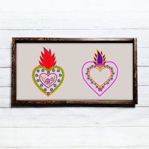 May include: Two framed embroidered heart designs with floral details. The hearts are in a traditional Mexican style with a red and purple flame on top. The hearts are surrounded by a border of flowers and beads.