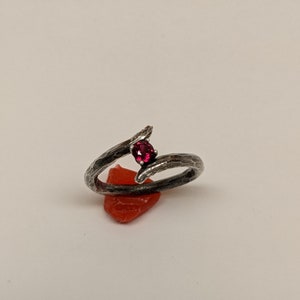 May include: A silver ring with a textured finish, featuring a single red gemstone set in the center. The ring is designed in a twisted branch shape.