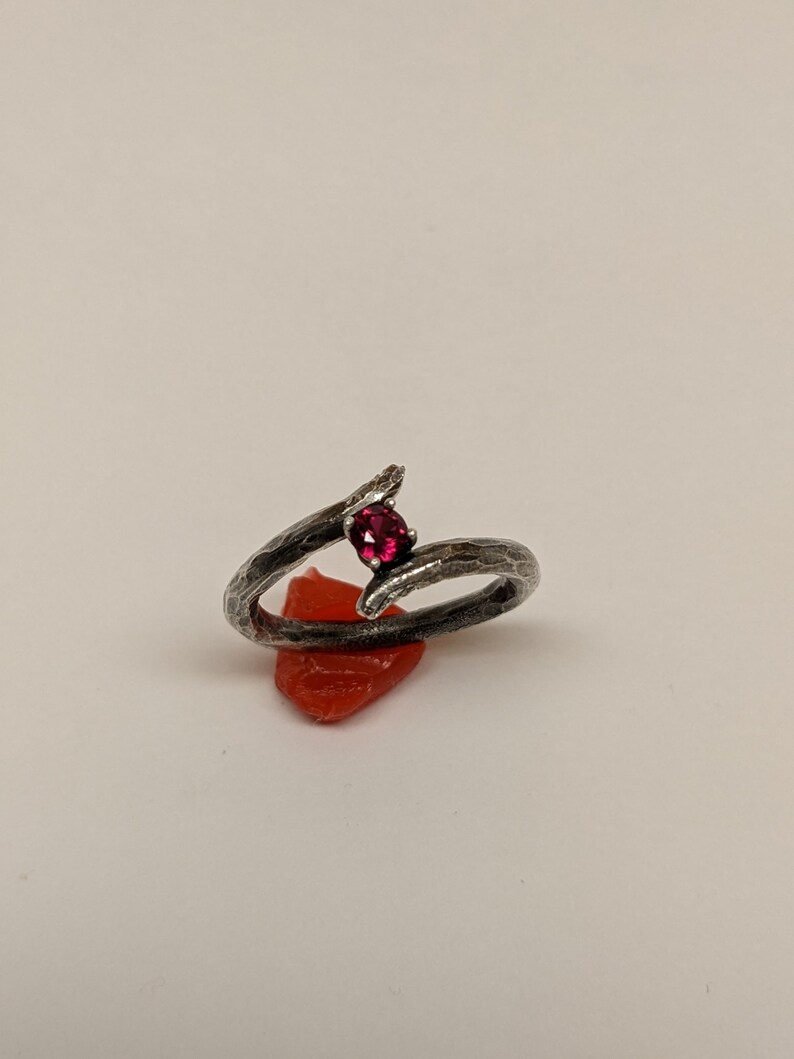 May include: A silver ring with a textured finish, featuring a single red gemstone set in the center. The ring is designed in a twisted branch shape.