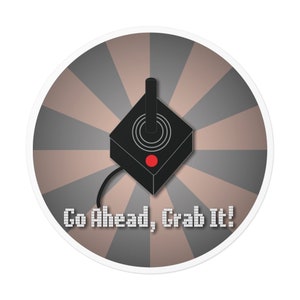 May include: A round sticker with a retro-style joystick in the center. The joystick is black with a red button and is surrounded by a gray and brown radial pattern. The text "Go Ahead, Grab It!" is printed in a pixelated font below the joystick.
