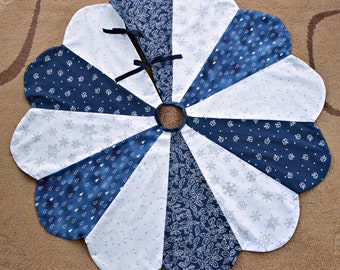 May include: A blue and white Christmas tree skirt with a snowflake, holly, and tree design. The skirt is made of fabric and has a scalloped edge.