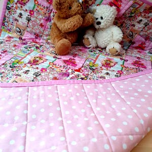 May include: A pink and white baby quilt with a white polka dot pattern on the back. The front of the quilt is made of patchwork fabric with a floral design and features two teddy bears.