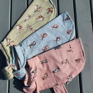 May include: Three quilted pet coats in pastel shades: pale green, light blue, and pink. Each coat features a repeating pattern of dog illustrations. The coats are designed with a curved shape and a folded collar.