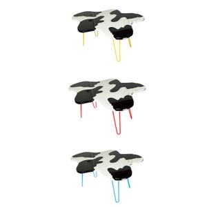 May include: Three black and white cow print coffee tables with different coloured metal legs. The first table has yellow legs, the second has red legs, and the third has blue legs.
