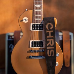 May include: A gold-colored electric guitar with a black strap that reads "CHRIS" in large, bold letters. The guitar has black pickups and silver hardware. The background is a warm, neutral tone.