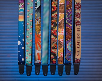 May include: A collection of eight guitar straps with various designs. The straps feature patterns including flamingos, a tiger, donuts, and the text "WE CAN DO IT". Each strap has a black leather end.