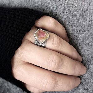 May include: A silver ring with a large, irregularly shaped pink and white gemstone. The ring's band has a textured design. The ring is worn on a finger, with a gray background.