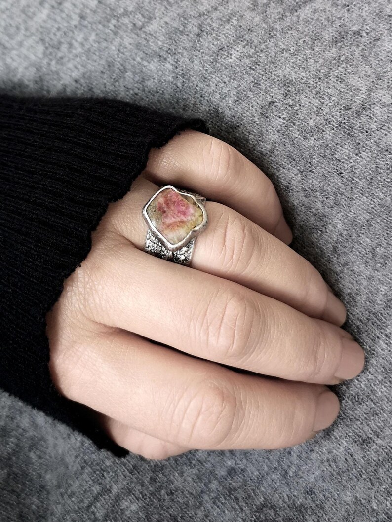 May include: A silver ring with a large, irregularly shaped pink and white gemstone. The ring's band has a textured design. The ring is worn on a finger, with a gray background.
