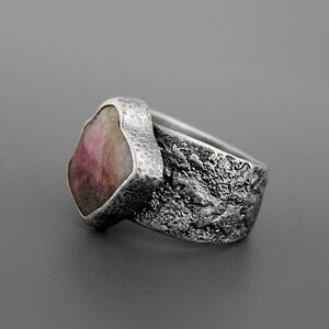 May include: A silver ring with a large, irregularly shaped pink gemstone. The band has a textured, organic design, and the setting around the stone has a hammered appearance. The ring is set against a gray background, highlighting its unique, handcrafted aesthetic.