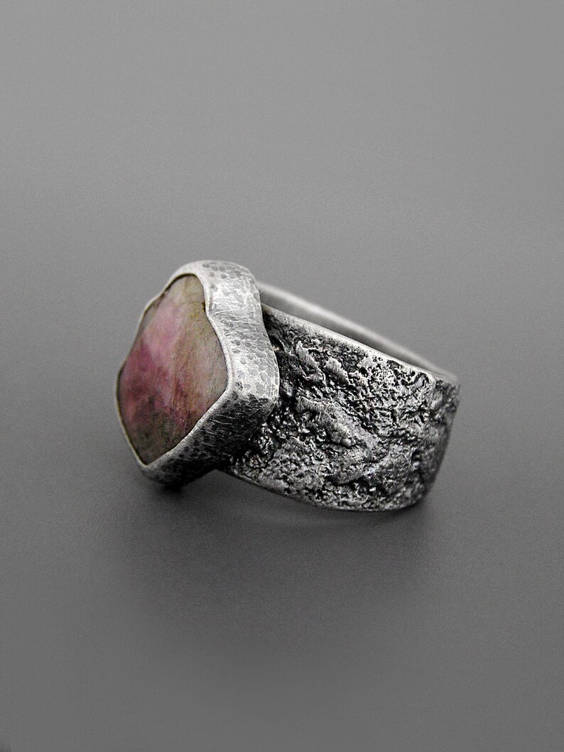 May include: A silver ring with a large, irregularly shaped pink gemstone. The band has a textured, organic design, and the setting around the stone has a hammered appearance. The ring is set against a gray background, highlighting its unique, handcrafted aesthetic.