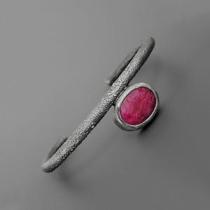 Puede incluir: Brazalete de puño plateado con una banda texturizada y una gema ovalada de color rosa intenso. La gema está engastada en un bisel plateado. El brazalete tiene un diseño abierto. Joyería moderna.