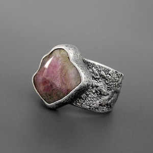 May include: A silver ring with a large, irregularly shaped pink gemstone. The band has a textured, hammered appearance. The gemstone is a light pink color with some darker inclusions.