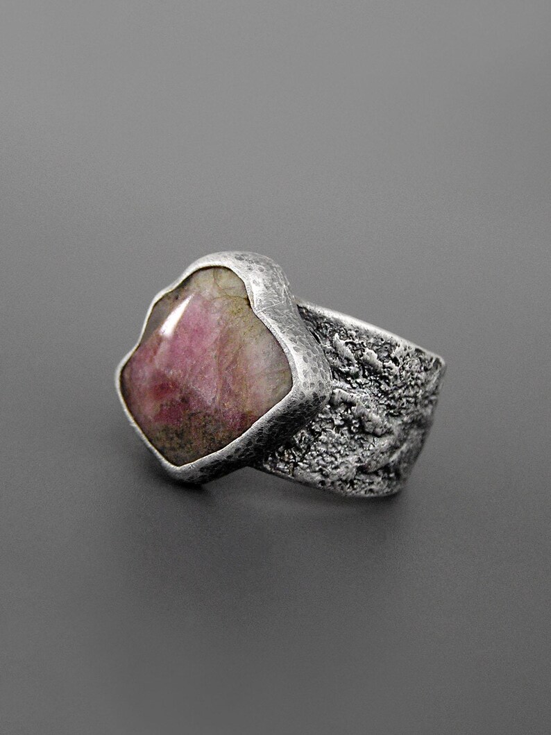 May include: A silver ring with a large, irregularly shaped pink gemstone. The band has a textured, hammered appearance. The gemstone is a light pink color with some darker inclusions.