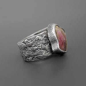 May include: A textured silver ring with a heart-shaped pink gemstone. The ring has a rough, organic texture on the band and a smooth, hammered bezel setting for the stone. The gemstone has a mix of pink and white hues.