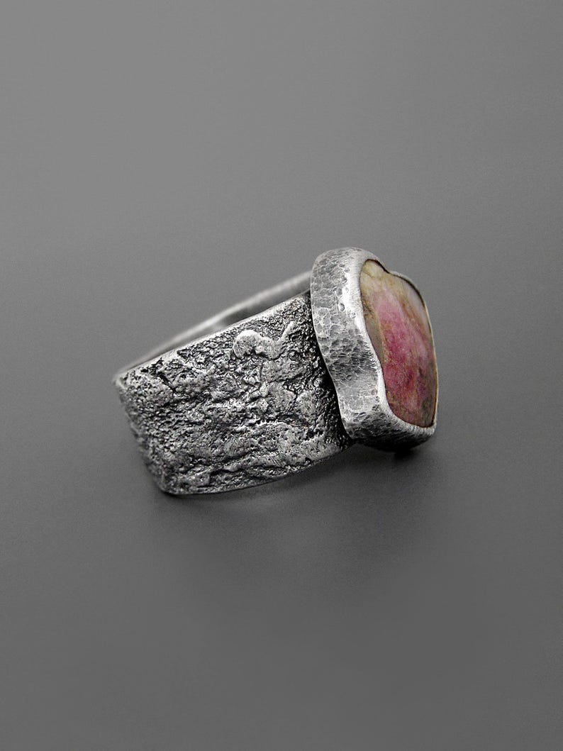 May include: A textured silver ring with a heart-shaped pink gemstone. The ring has a rough, organic texture on the band and a smooth, hammered bezel setting for the stone. The gemstone has a mix of pink and white hues.