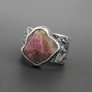 May include: A silver ring featuring a large, irregularly shaped pink gemstone. The stone is set in a textured silver band. The ring's design is bold and eye-catching, with a rustic, handcrafted aesthetic. The gemstone has a mix of pink and green hues.