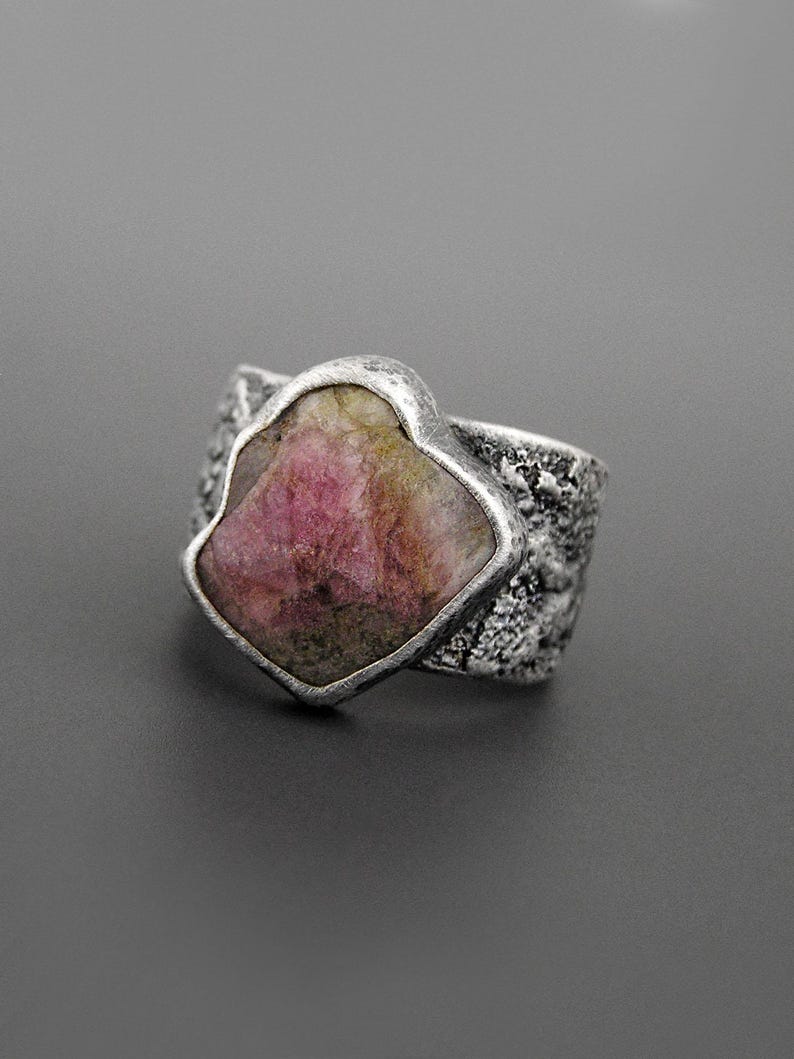 May include: A silver ring featuring a large, irregularly shaped pink gemstone. The stone is set in a textured silver band. The ring's design is bold and eye-catching, with a rustic, handcrafted aesthetic. The gemstone has a mix of pink and green hues.