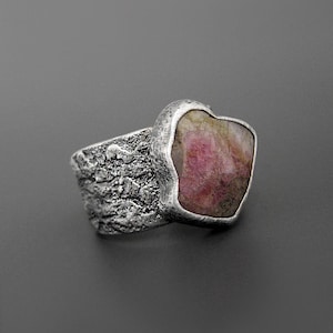 May include: A silver ring with a textured band and a large, irregularly shaped pink gemstone. The gemstone is set in a smooth bezel, contrasting with the rough texture of the band. The ring is displayed against a gray background.