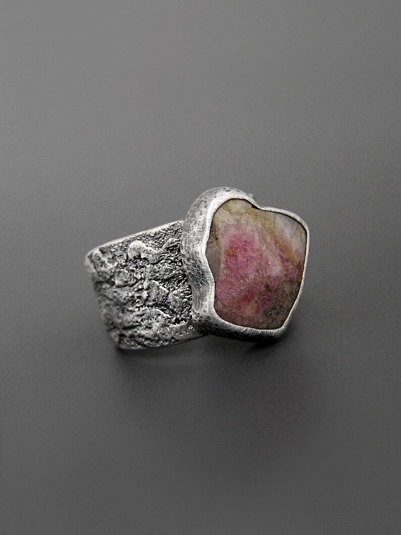 May include: A silver ring with a textured band and a large, irregularly shaped pink gemstone. The gemstone is set in a smooth bezel, contrasting with the rough texture of the band. The ring is displayed against a gray background.