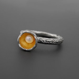 May include: A silver ring with a textured band and a gold-colored interior setting. The setting holds a small, round, white pearl. The ring's design is organic and elegant, suitable for everyday wear or special occasions.