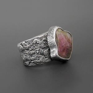 May include: A silver ring with a textured band and a large, irregular pink gemstone. The gemstone is set in a smooth, silver bezel, contrasting with the rough texture of the band. The ring has a rustic, handcrafted appearance.
