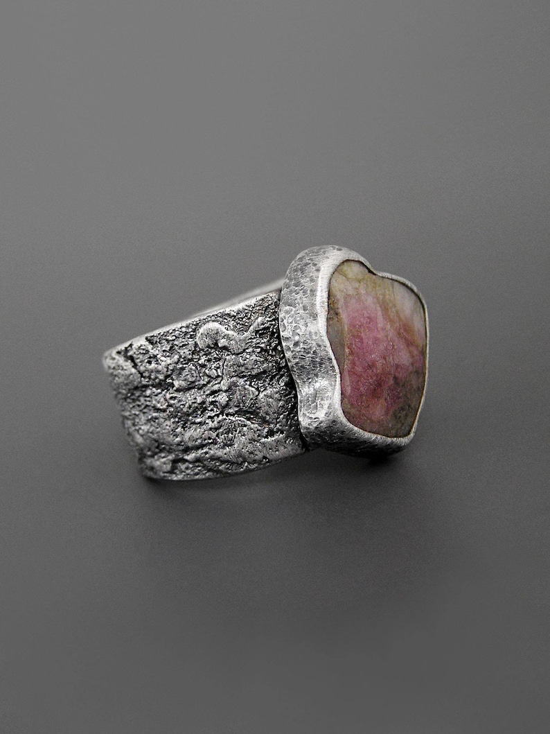 May include: A silver ring with a textured band and a large, irregular pink gemstone. The gemstone is set in a smooth, silver bezel, contrasting with the rough texture of the band. The ring has a rustic, handcrafted appearance.