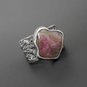 May include: A silver ring featuring a large, irregularly shaped pink gemstone. The ring's band has a textured, organic design, and the gemstone is set in a smooth, silver bezel. The overall aesthetic is rustic and natural, suitable for everyday wear.