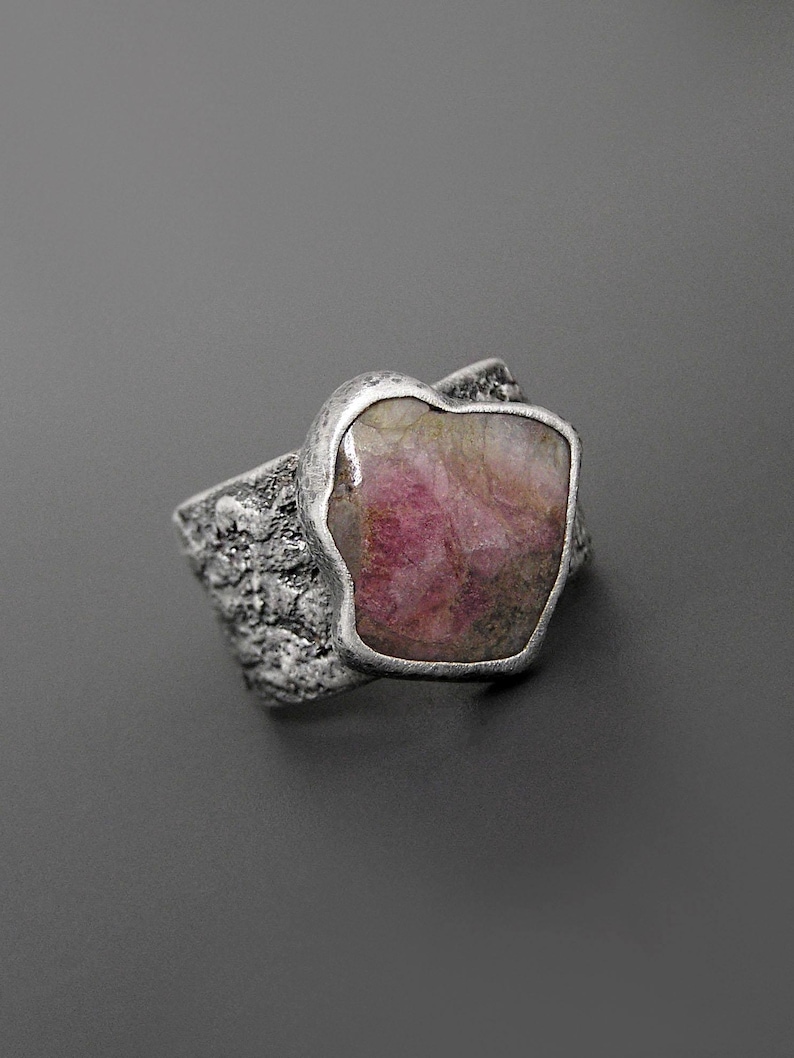 May include: A silver ring featuring a large, irregularly shaped pink gemstone. The ring's band has a textured, organic design, and the gemstone is set in a smooth, silver bezel. The overall aesthetic is rustic and natural, suitable for everyday wear.