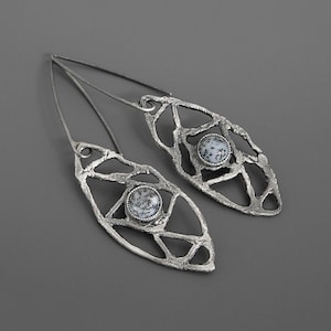 May include: A pair of silver-toned dangle earrings. Each earring features an openwork, leaf-shaped design with a central, round stone. The stones have a speckled, black and white pattern. The earrings have a long, thin hook.