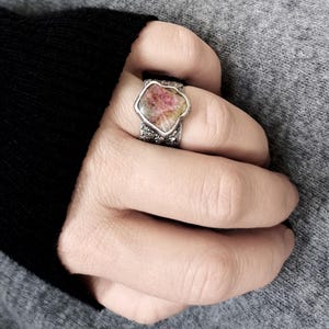 May include: A silver ring featuring a large, irregularly shaped gemstone with pink and yellow hues. The ring's band has a textured, possibly oxidized, appearance. The gemstone is the focal point, set in a bezel setting.