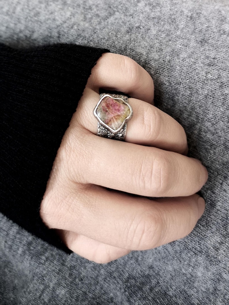 May include: A silver ring featuring a large, irregularly shaped gemstone with pink and yellow hues. The ring's band has a textured, possibly oxidized, appearance. The gemstone is the focal point, set in a bezel setting.