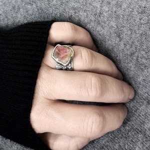 May include: A silver ring with a large, irregularly shaped gemstone. The gemstone has a pink and yellow color palette. The ring's band has a textured, detailed design. The ring is worn on a hand, with a gray sweater in the background.