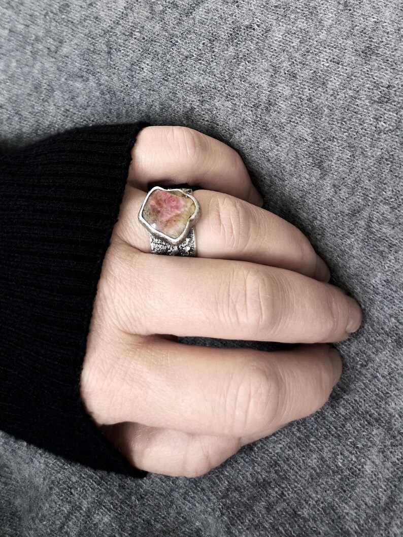 May include: A silver ring with a large, irregularly shaped gemstone. The gemstone has a pink and yellow color palette. The ring's band has a textured, detailed design. The ring is worn on a hand, with a gray sweater in the background.