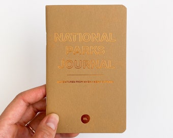 May include: A brown journal with gold foil lettering that reads "National Parks Journal" and "Adventures from when I went outside". The journal has a small gold circle with the letter "m" in the lower right corner.