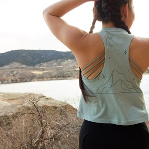 May include: A woman wearing a light blue tank top with a mountain and moon graphic and black athletic shorts. The tank top has a racerback style with multiple straps.