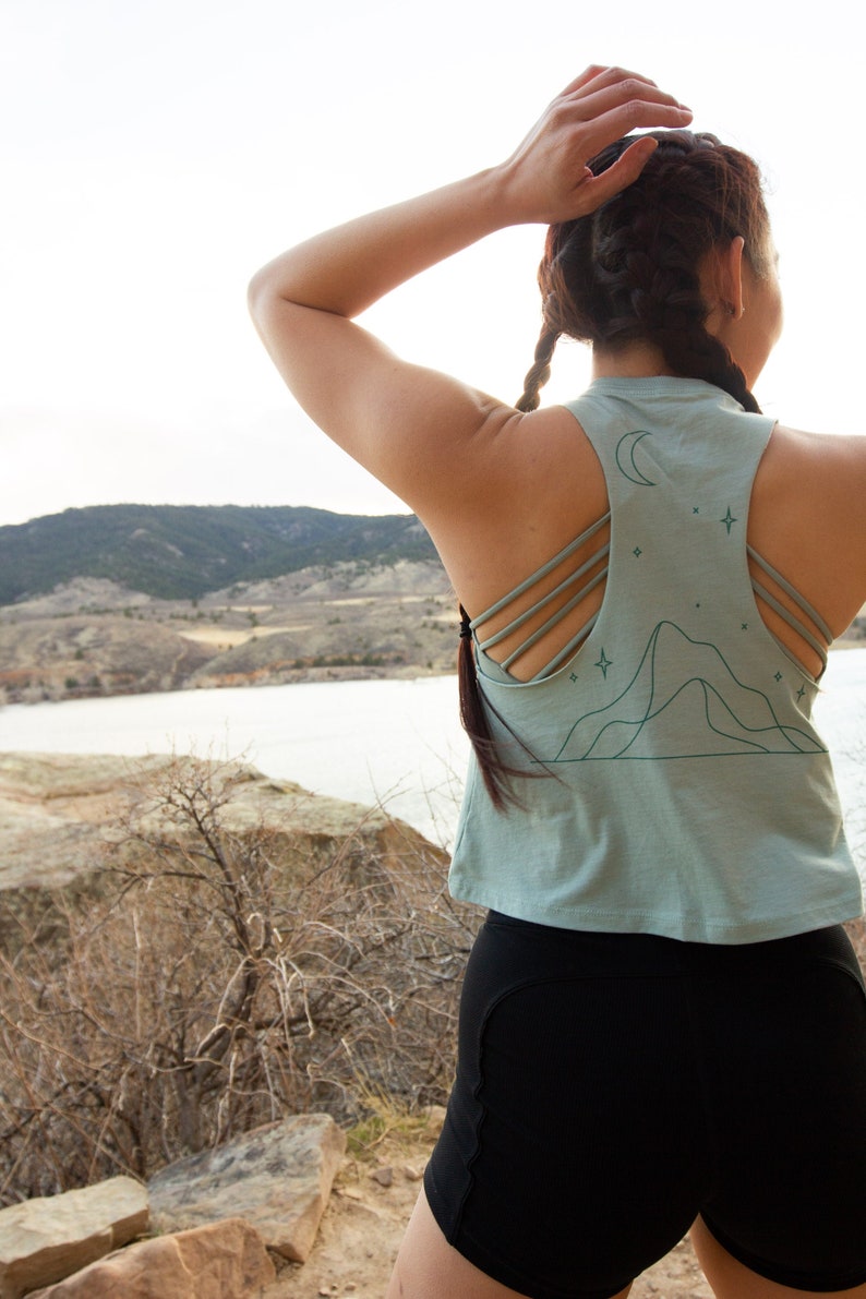 May include: A woman wearing a light blue tank top with a mountain and moon graphic and black athletic shorts. The tank top has a racerback style with multiple straps.