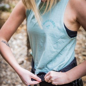 May include: A woman wearing a light blue tank top with the text "MONOPKT." in a blue arc with two stars above the text. She is adjusting a black climbing harness with a blue and orange strap.