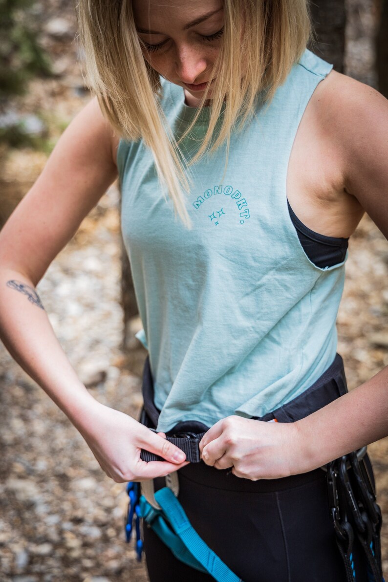 May include: A woman wearing a light blue tank top with the text "MONOPKT." in a blue arc with two stars above the text. She is adjusting a black climbing harness with a blue and orange strap.
