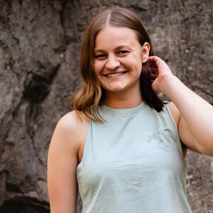 May include: A woman with long brown hair is smiling at the camera. She is wearing a light blue sleeveless shirt with the text "MONOKINI" printed on the front. The shirt has a star design.