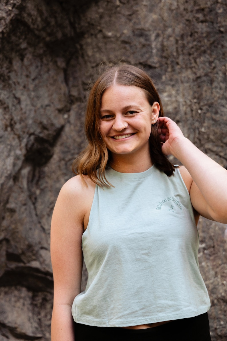 May include: A woman with long brown hair is smiling at the camera. She is wearing a light blue sleeveless shirt with the text "MONOKINI" printed on the front. The shirt has a star design.