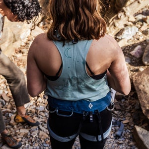 May include: A woman wearing a blue and green tank top with a moon and stars design. She is wearing a black harness with a blue belt and black pants.