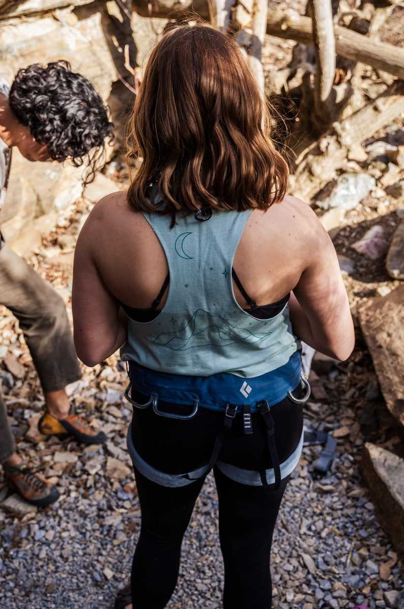May include: A woman wearing a blue and green tank top with a moon and stars design. She is wearing a black harness with a blue belt and black pants.