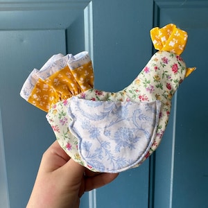 May include: A handmade fabric chicken with a floral pattern in shades of pink, green, and white. The chicken has a yellow comb and tail feathers. A blue and white floral pocket is on the front.