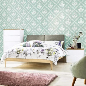 May include: A white bed with a floral patterned duvet cover and a light gray headboard. The bed is in a bedroom with a light green damask patterned wallpaper, a white dresser, and a light gray chair.