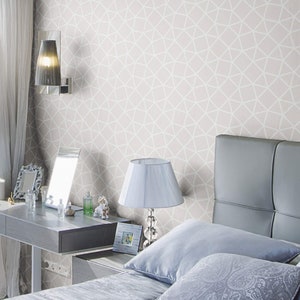 May include: A bedroom with a gray headboard, a white lamp with a white lampshade, and a white geometric patterned wallpaper. The bed has a blue and gray patterned pillow and a white pillow.