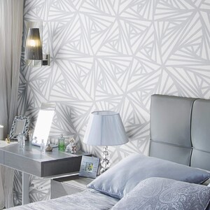 May include: A bedroom with a gray headboard and a white and gray geometric patterned wallpaper. There is a bedside table with a lamp and a picture frame.