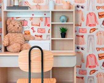 May include: A light wood desk with a brown teddy bear sitting on the surface. The desk has a light wood chair with a black metal frame. The wall behind the desk is covered in a wallpaper with a pattern of pink and white clothing items, including dresses, shirts, pants, and purses.