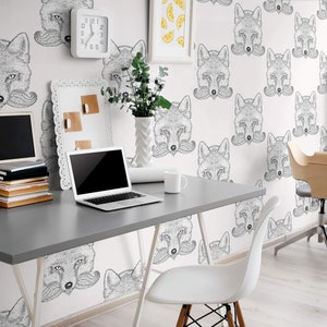 May include: A white desk with a laptop, a white mug, and a stack of books. The desk is in front of a white wall with a wallpaper pattern of fox faces with mustaches. There is a white chair in the foreground and a wooden chair in the background.